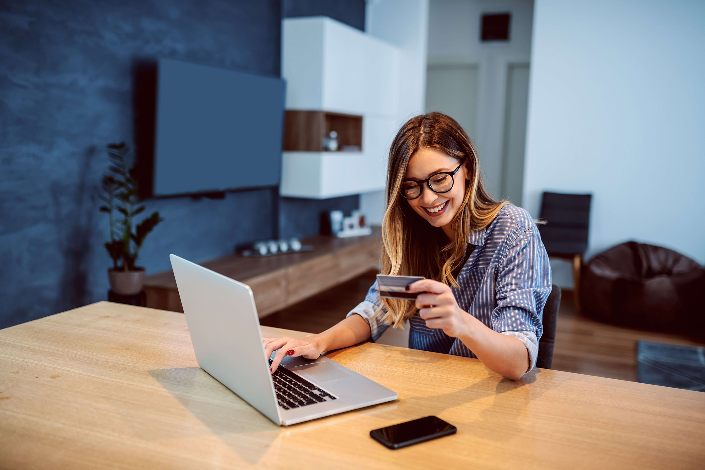 Mujer sonriente usando una notebook en una mesa, sosteniendo una tarjeta de crédito para realizar una compra online en un entorno hogareño moderno.